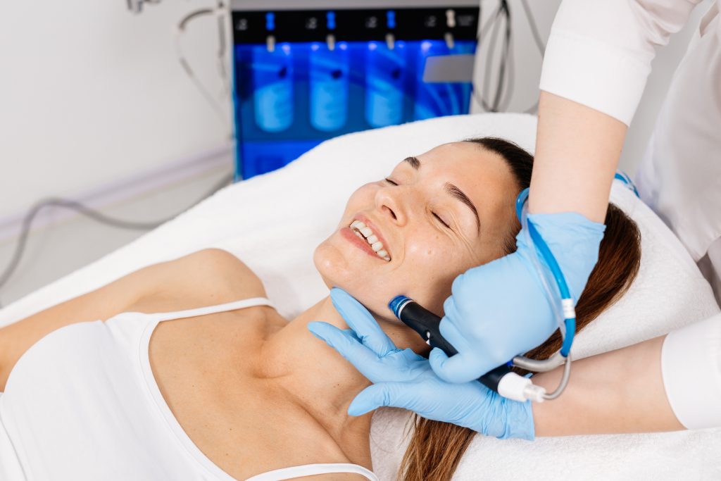 Orta yaşlı bir kadın profesyonel bir güzellik salonunda yüz bakımı yaptırıyor. Yakın çekim bir güzellik uzmanı yüz cildini temizlemek ve gençleştirmek için bir hidropeeling cihazını kullanıyor.